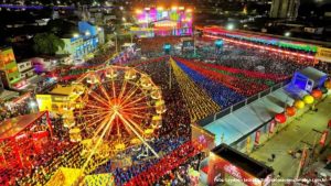 Uma vista aérea noturna vibrante do Parque do Povo em Campina Grande durante o São João, com uma roda-gigante iluminada no primeiro plano à esquerda. O parque está repleto de pessoas e barracas, com longas faixas de luzes coloridas estendendo-se pelo chão, criando um efeito visual impressionante. Ao fundo, um palco grande e iluminado com luzes de show e telões. A cena está cheia de energia e celebração.